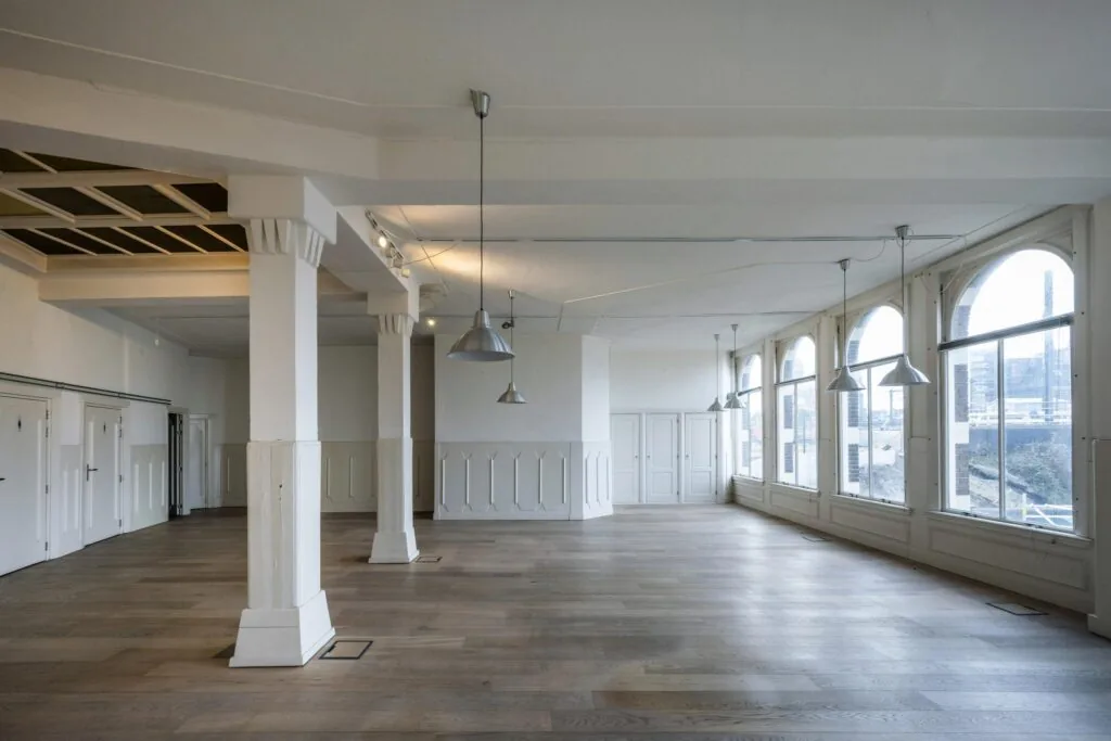 Lichte, ruime interieurfoto van een leegstaand pand aan de De Ruijterkade met houten vloer, witte pilaren en grote boogramen.